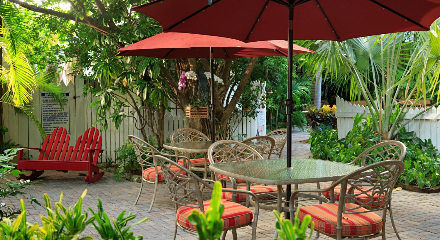 Patio area with square tables and umbrellas with red cushioned chairs surrounded by dense greenery and red wooden slated seat