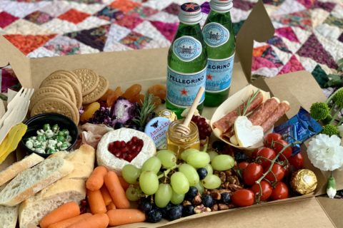 Box on patchwork quilt with 2 green bottles of Pellegrino mineral water and assortment of fruits, vegetables and snacks for picnic.