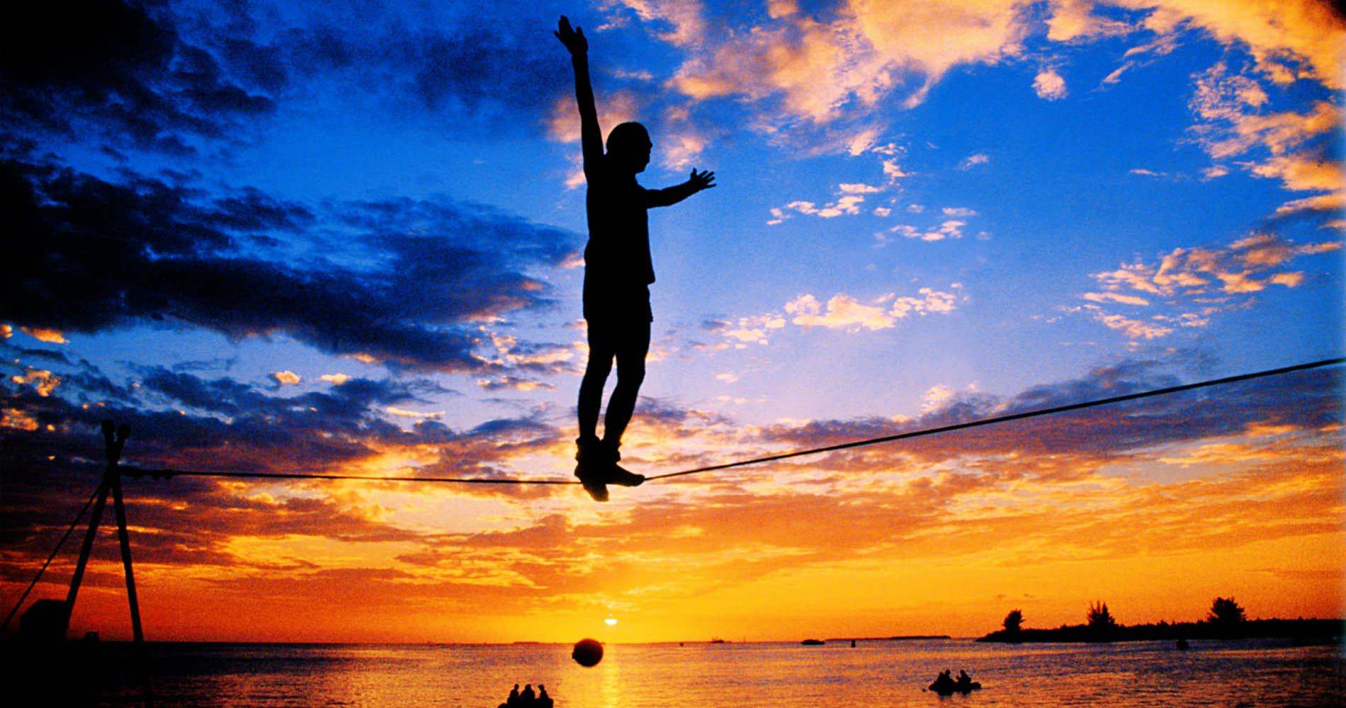 Silhouette figure standing on tightrope over the ocean with sun setting in background