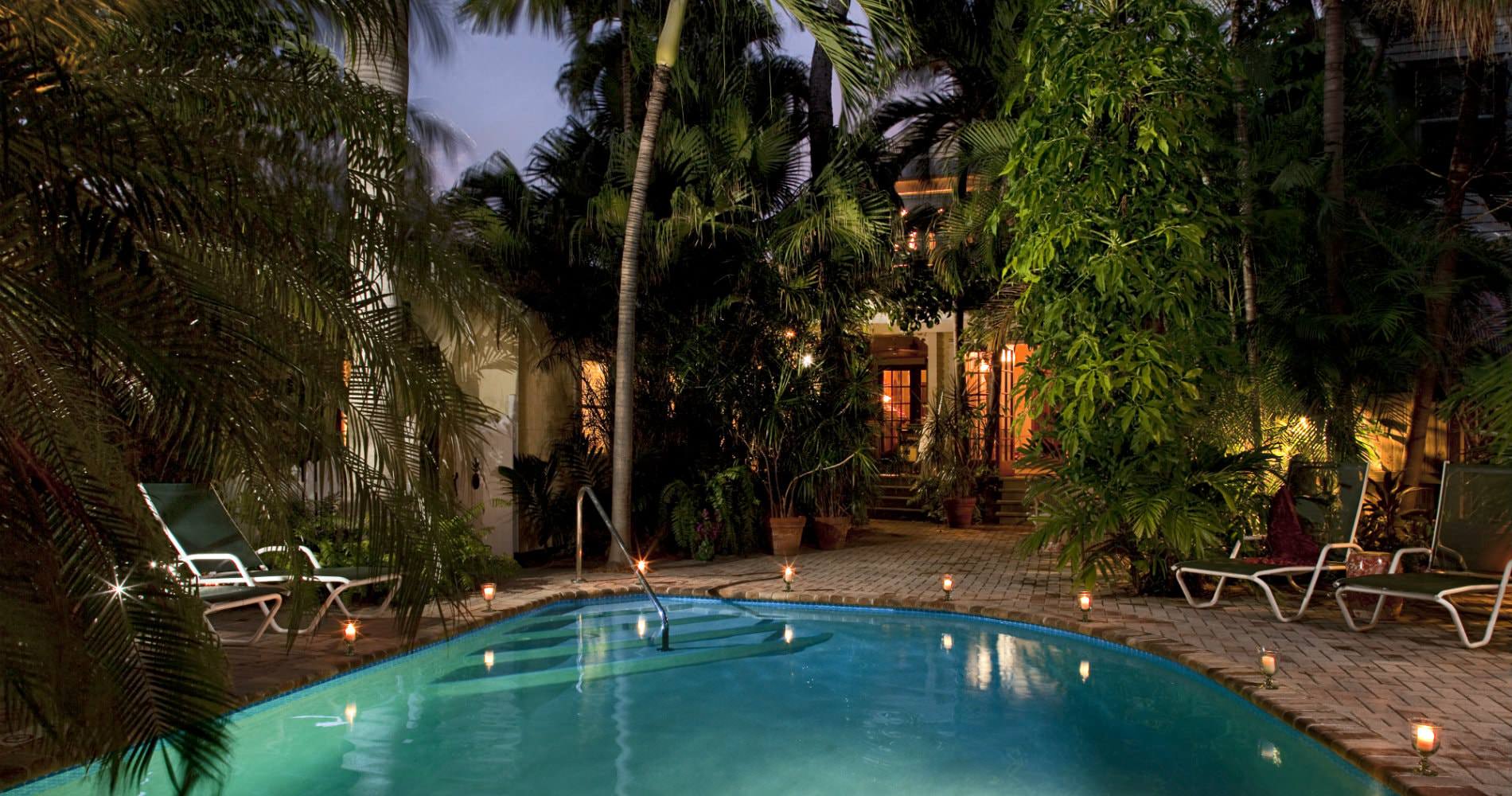 Oblong pool surrounded by trees, lounge chairs and lighting on deck at night