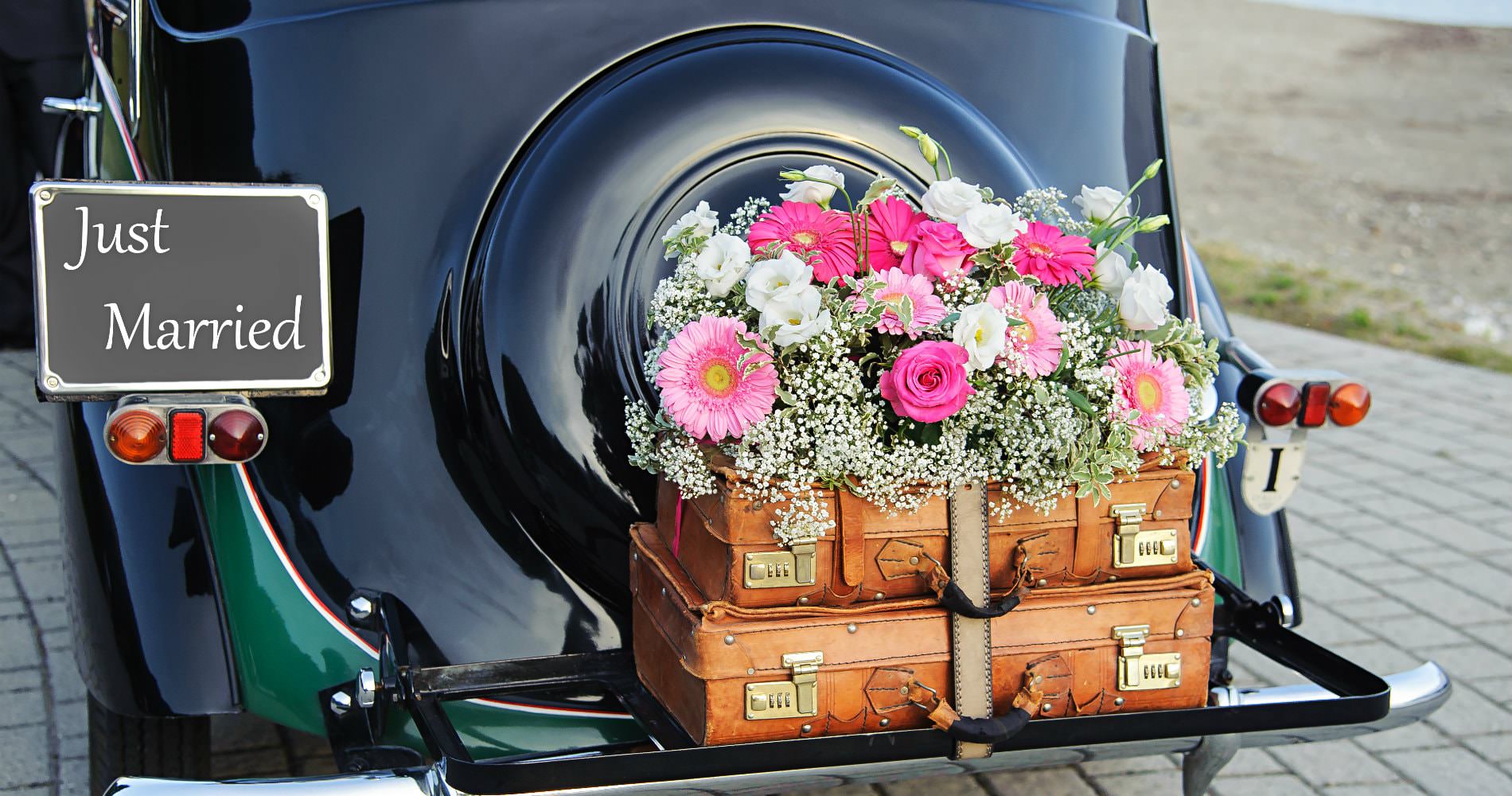 Shiny black antique car with brown trunks, fresh pink flowers, and a Just Married sign on the tailgate