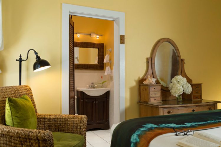 Yellow bedroom with antique dresser with mirror with connecting bathroom with slatted door and wicker chair