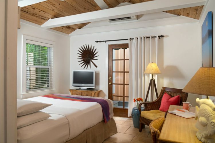 Bedroom with window with mini blinds with dresser and television on wood table at end of bed and wicker chair