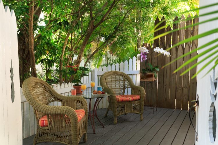 Wood deck with cushioned wicker chairs and round glass table surrounded by white and brown picket fence