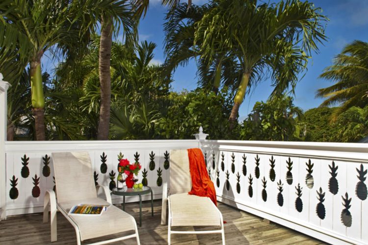 Enclosed patio with cream lounge chairs and glass table surrounded by dense greenery and orange towel draped over 1 chair