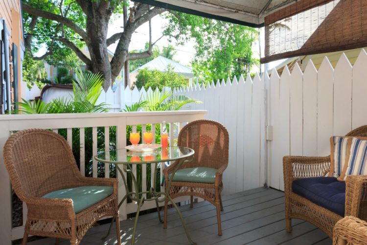 Covered patio with round glass table with cushioned wicker chairs surrounded by white picket fence