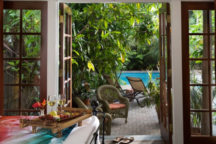 View from bedroom leading to pool with lounge and wicker chairs surrounded by dense greenery