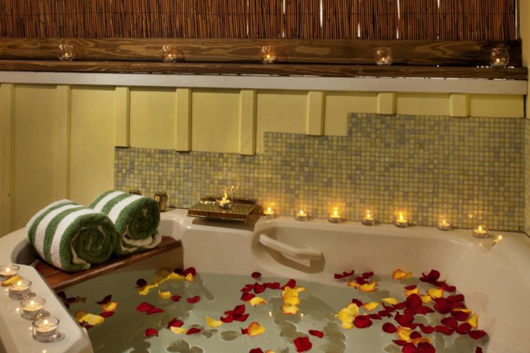 Large jacuzzi tub with red and yellow flower petals in water surrounded by small lit candles for mood lighting