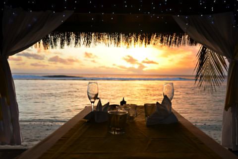 Straw hut with 2 wine glasses on table next to ocean with sun setting in background