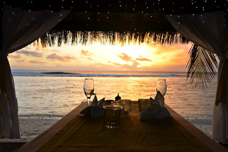 traw hut with 2 wine glasses on table next to ocean with sun setting in background