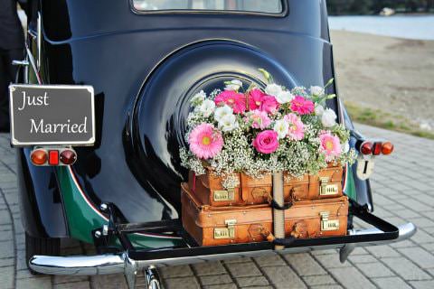 Shiny black antique car with brown trunks, fresh pink flowers, and a Just Married sign on the tailgate