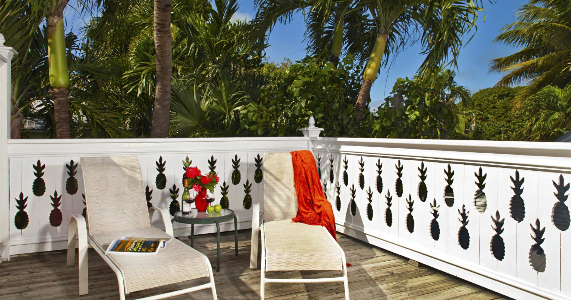 Enclosed patio with cream lounge chairs and glass table surrounded by dense greenery and orange towel draped over 1 chair