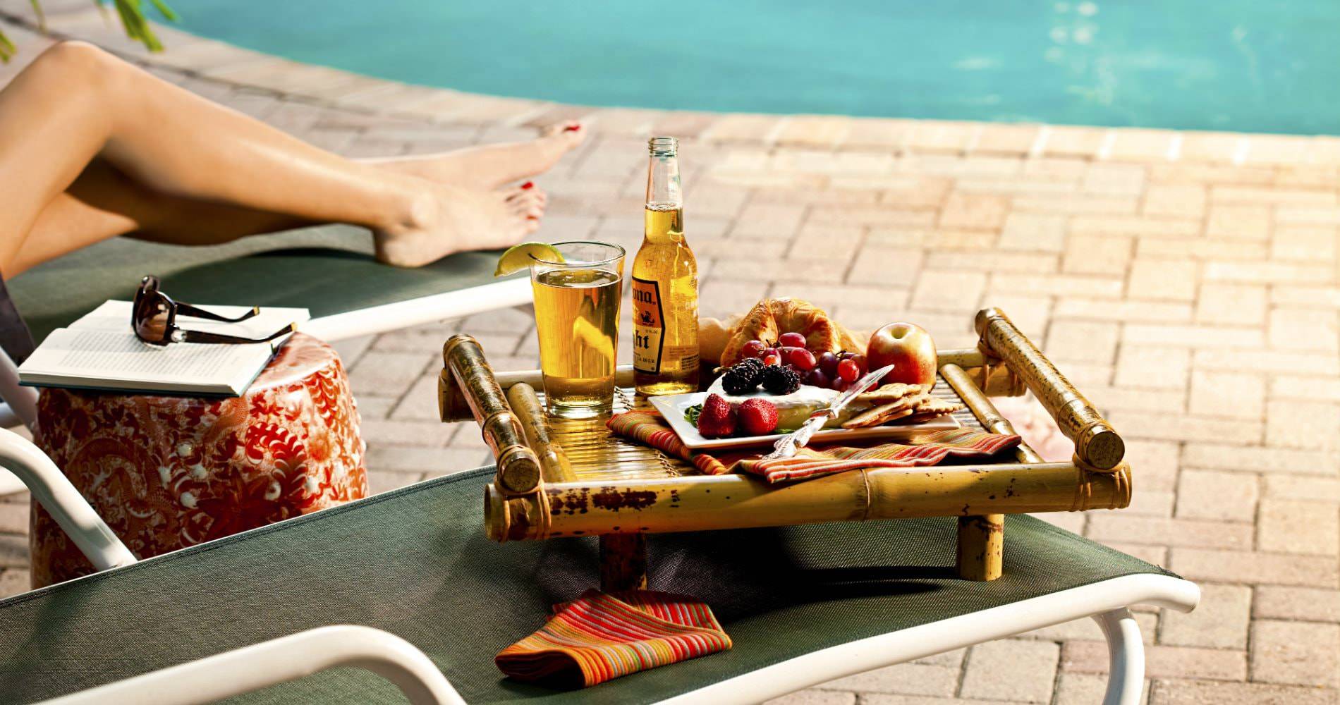 Woman laying on a lounge chair by the pool with a book, sunglasses and a try of beer and fruit