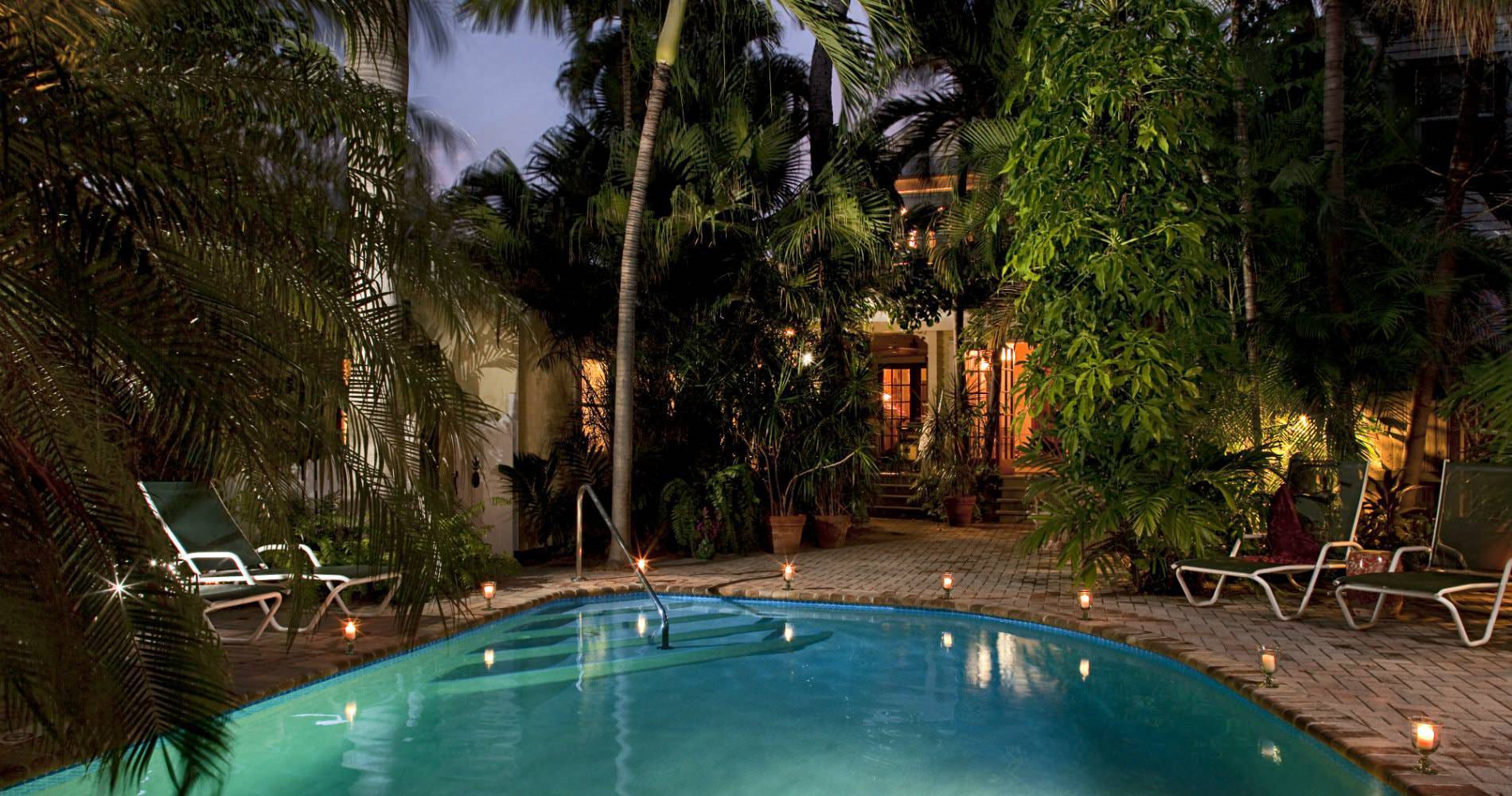 Oblong pool surrounded by trees, lounge chairs and lighting on deck at night